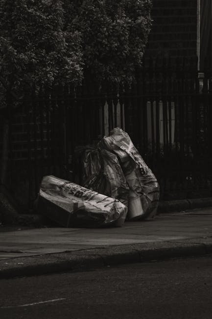 Four large black plastic garbage bags, some appearing crumpled and loosely tied, are piled on the pavement in front of a wooden fence with vertical slats. The bags contain household waste or rubbish,