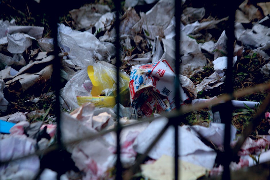 Four large black plastic garbage bags, some appearing crumpled and loosely tied, are piled on the pavement in front of a wooden fence with vertical slats. The bags contain household waste or rubbish,