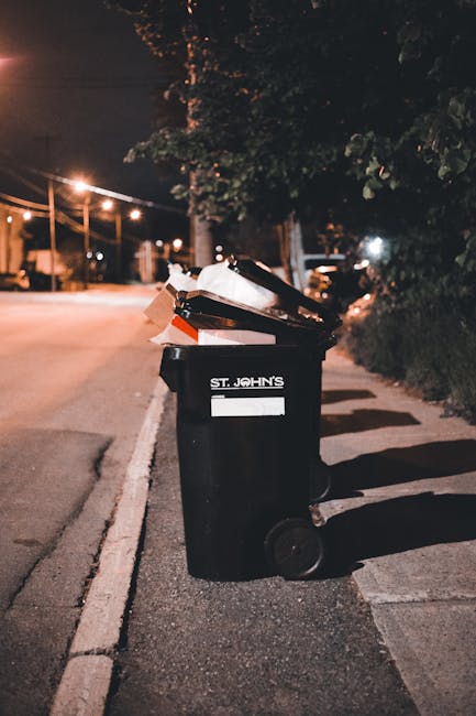 A large pile of mixed household waste and rubbish, consisting of crumpled cardboard boxes, plastic bags, paper waste, and discarded packaging, is overflowing from a grey recyclable waste bin with a bl